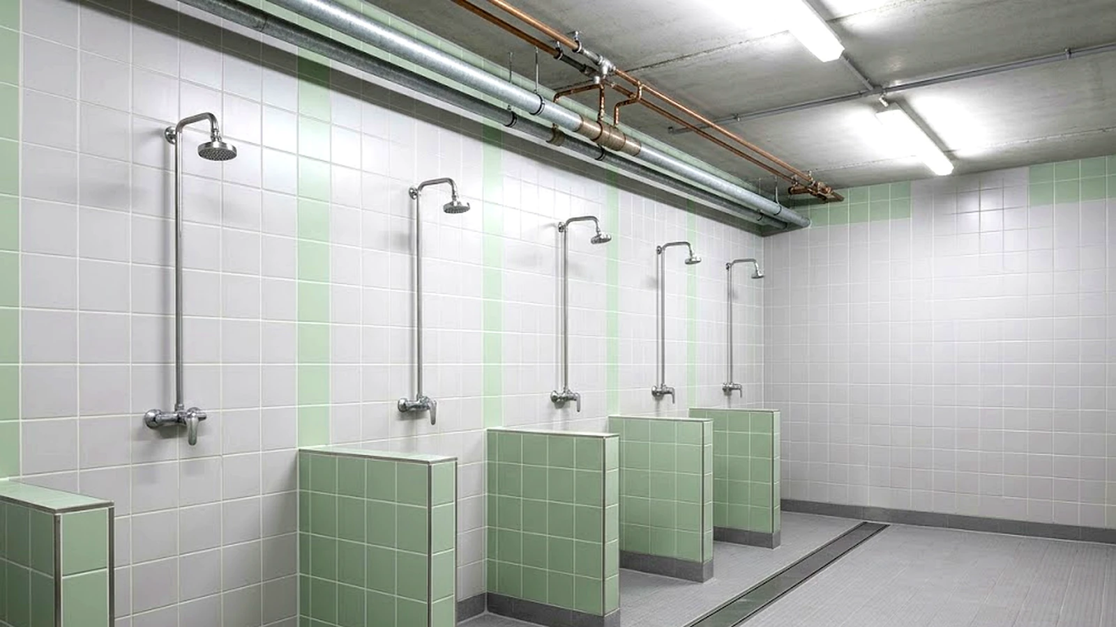 An empty communal shower room with a row of shower heads on a tiled wall, dry floor and exposed supply pipework along the ceiling.
